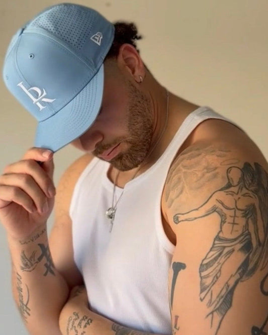 Man wearing a blue cap and white tank top with tattoos on arms, standing in a garage.