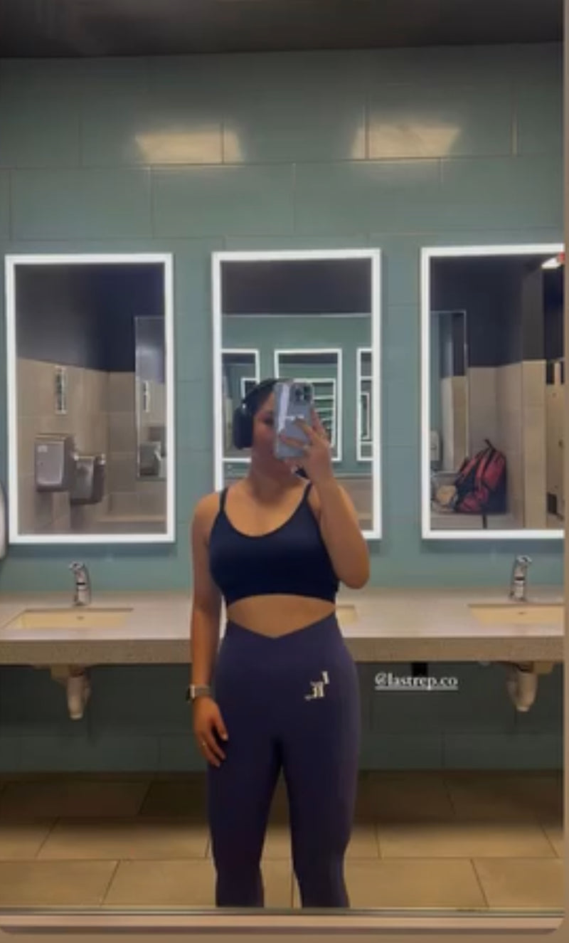 Person taking a mirror selfie in a bathroom wearing a black Last Rep sports bra and leggings.