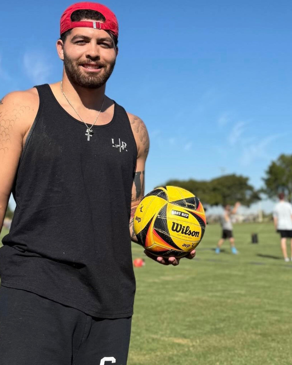 Man holding a volleyball sports ball on a grassy field with a clear blue sky, wearing black tank top