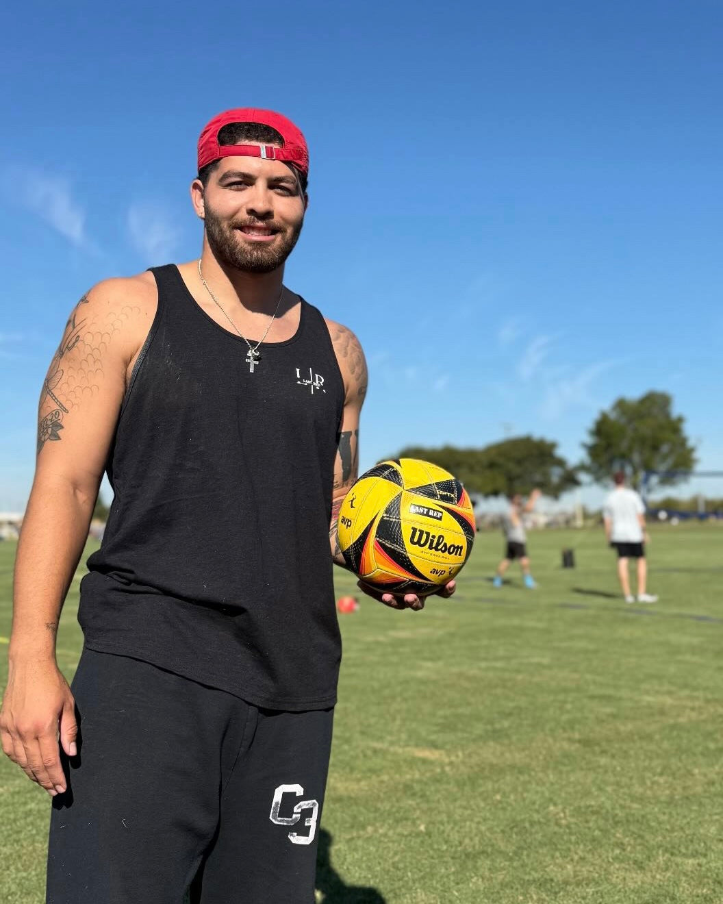 Man holding a volleyball sports ball on a grassy field with a clear blue sky, wearing black tank top