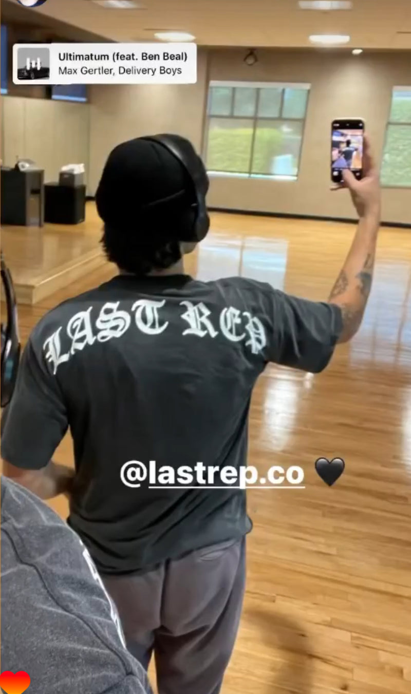Person wearing a black 'LAST REP' t-shirt in a dance studio, holding a phone.