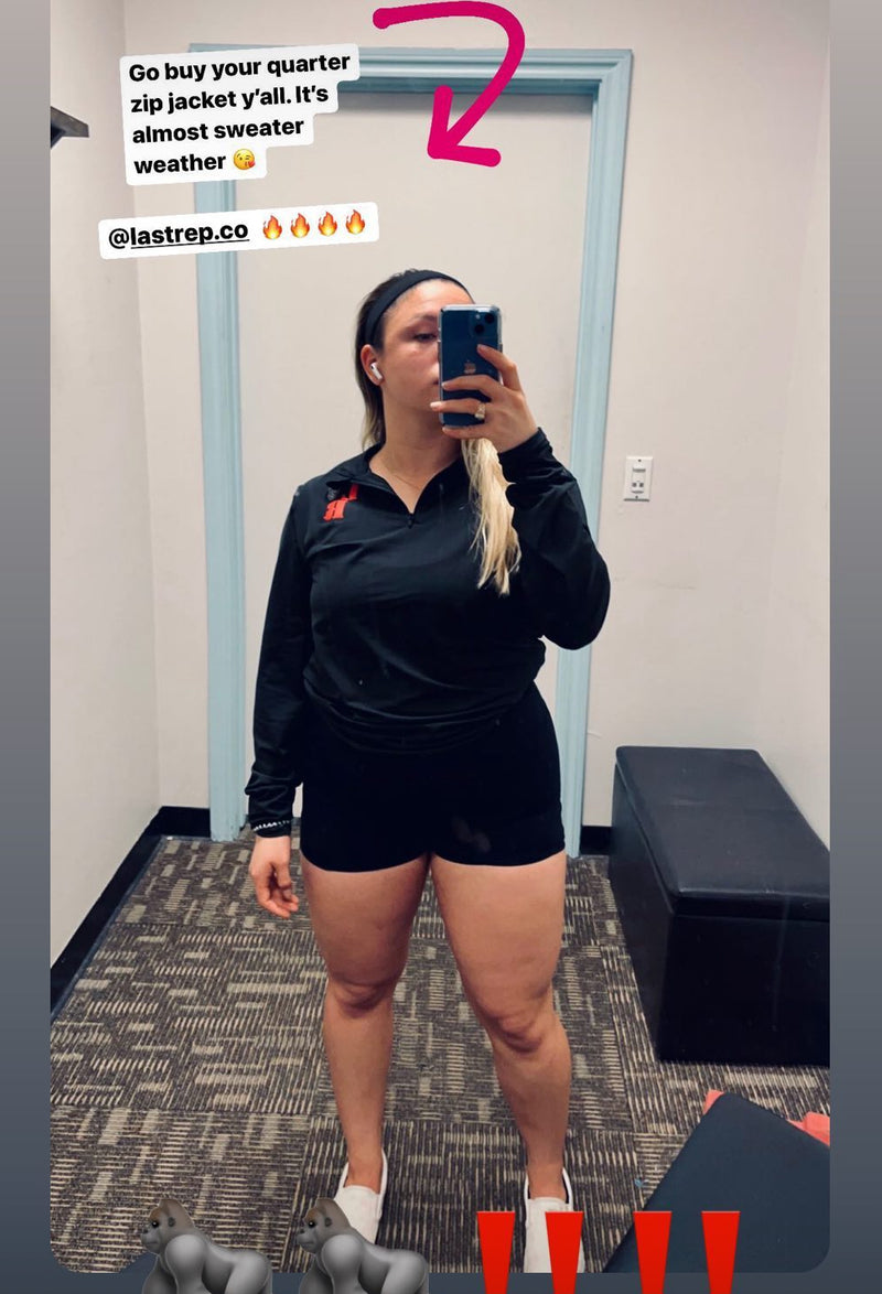 Person taking a mirror selfie wearing a black Last Rep quarter zip jacket and shorts indoors.