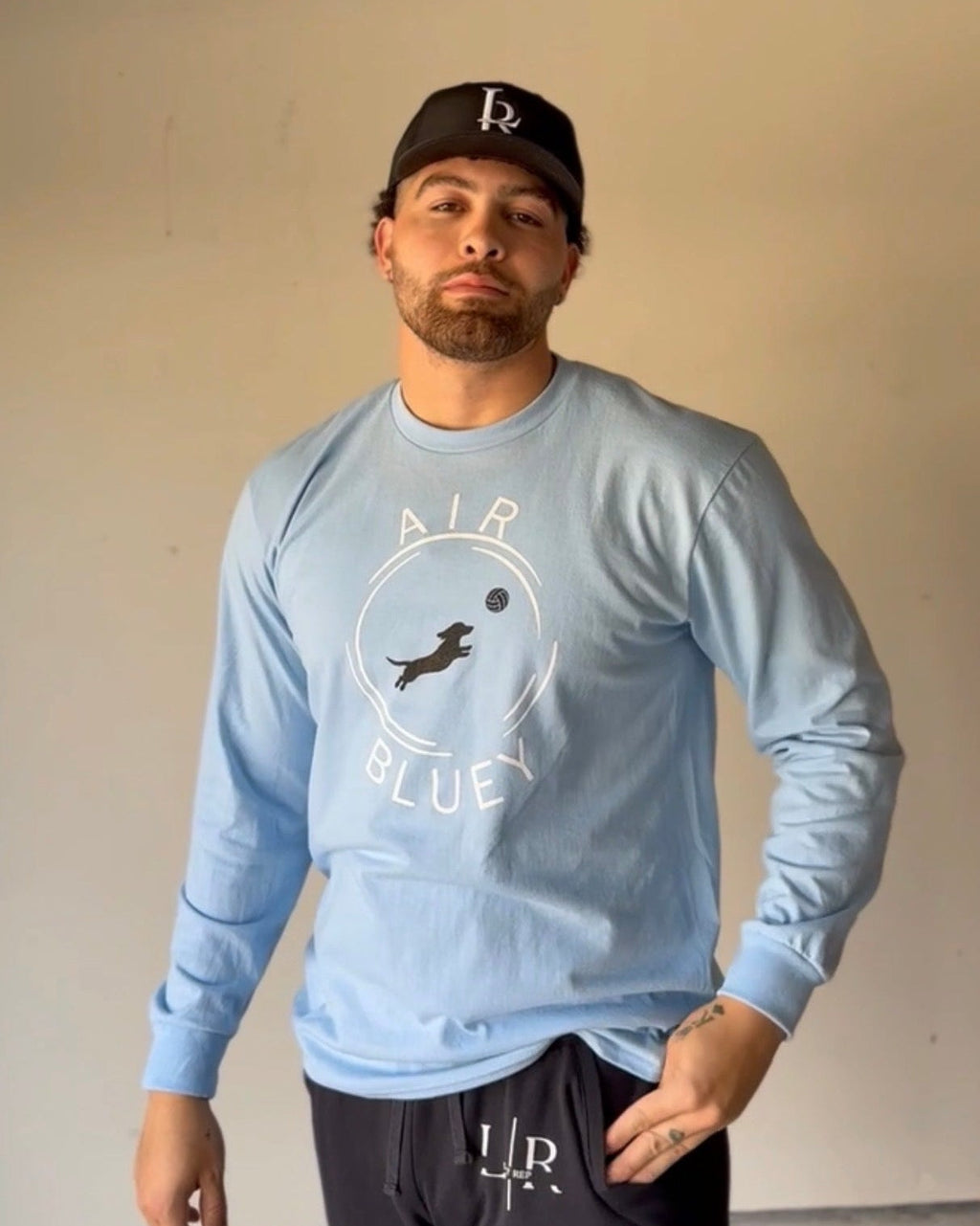 Man wearing a light blue long-sleeve shirt with a logo and dark pants against a plain background
