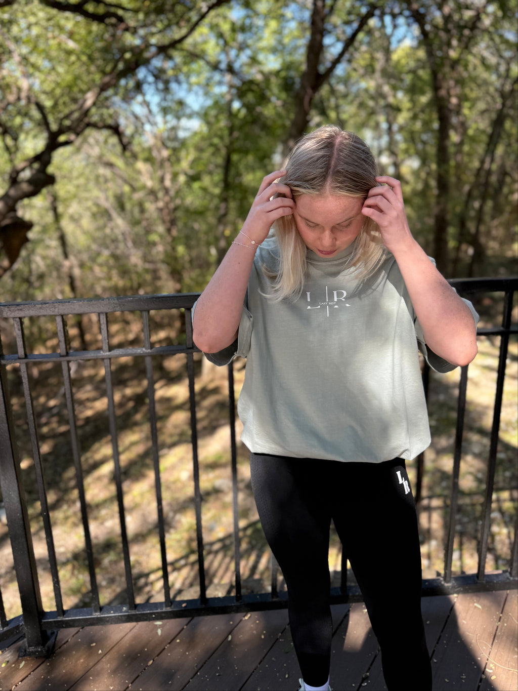 Person wearing a gray t-shirt with a logo on a wooden deck with trees in the background