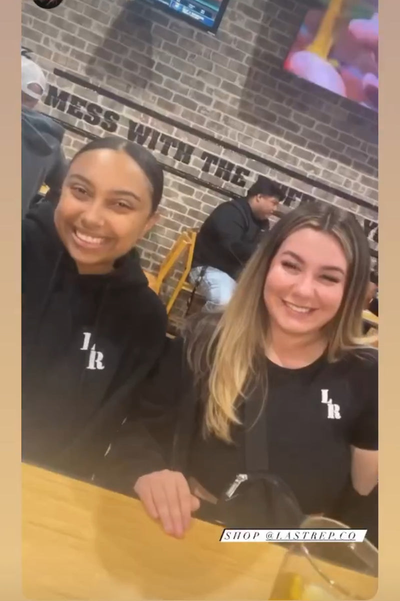 Two people wearing black Last Rep shirts with a logo, sitting at a table with a brick wall and text in the background.
