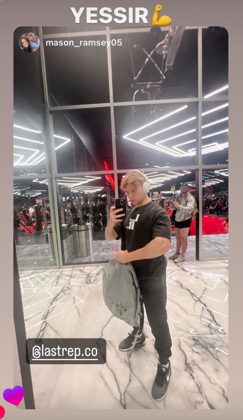 Person taking a mirror selfie in a gym with Last Rep black pump cover with gym equipment and people in the background.