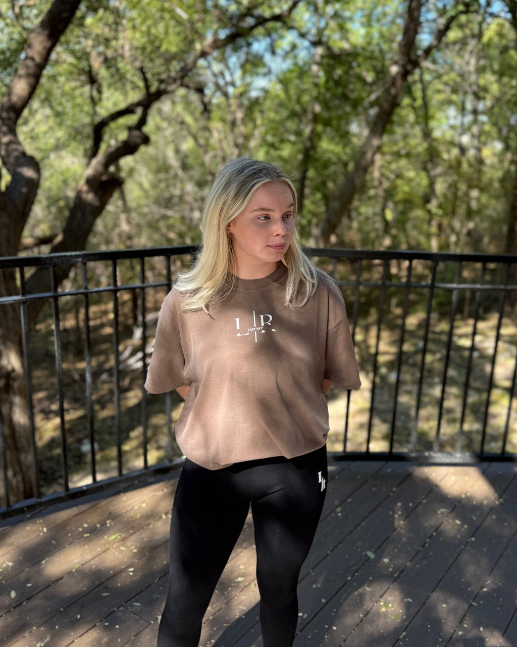 Person wearing a beige t-shirt with a logo, standing on a wooden deck with trees in the background