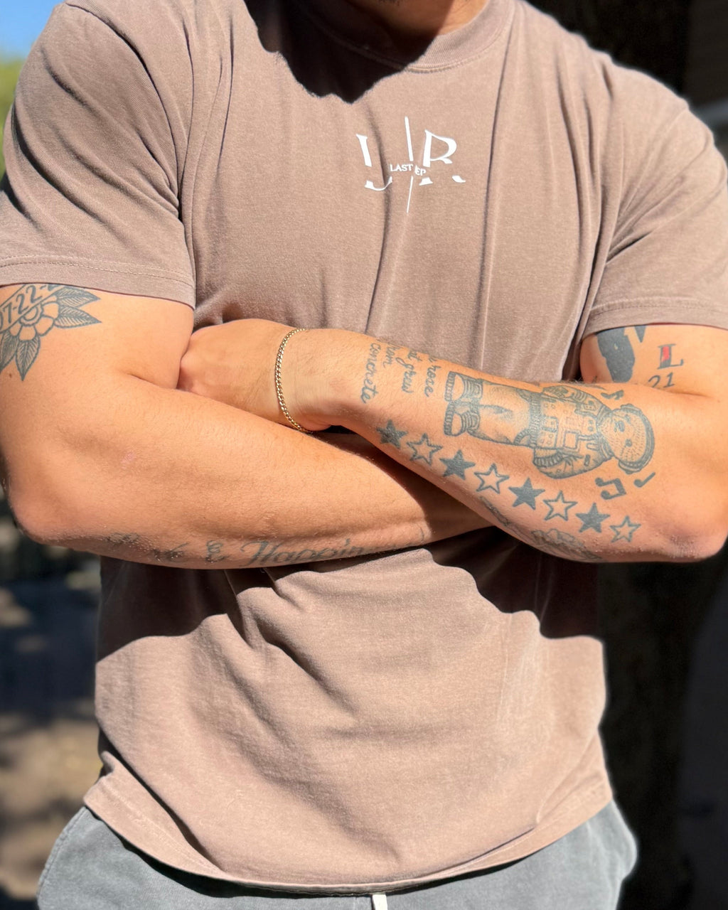 Person wearing a brown t-shirt with visible tattoos on arms
