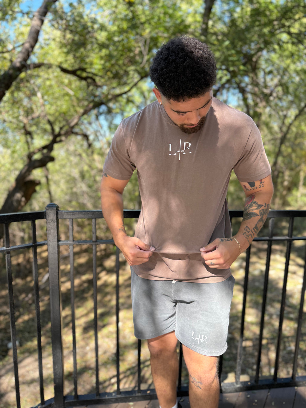 Man wearing a brown t-shirt and gray shorts with visible branding outdoors.