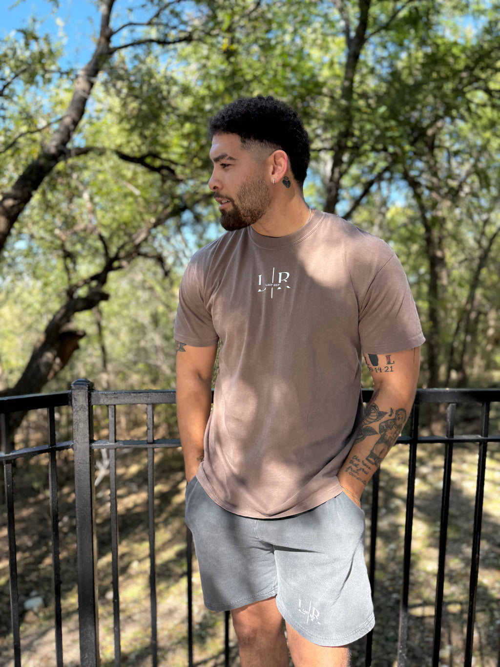 Man wearing a brown t-shirt with a logo and gray shorts standing outdoors.