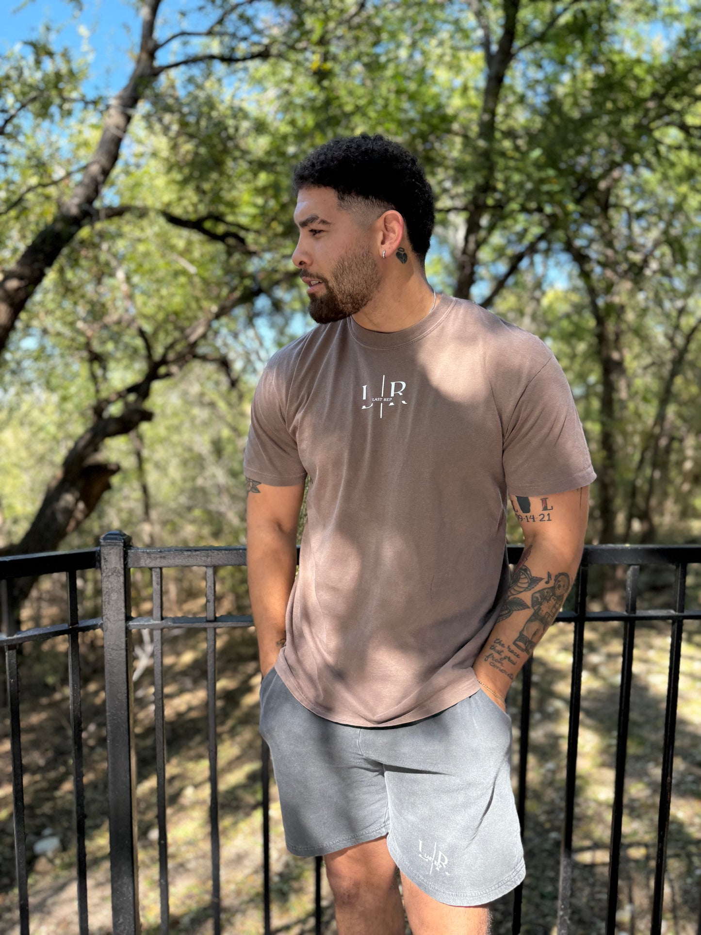 Man wearing a brown t-shirt with a logo and gray shorts standing outdoors.