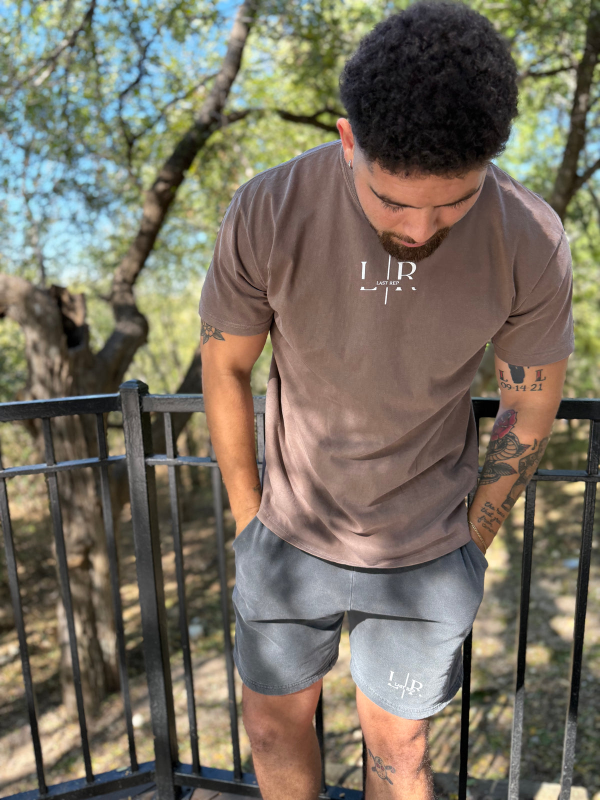 Man wearing a brown t-shirt and gray shorts with a logo outdoors.