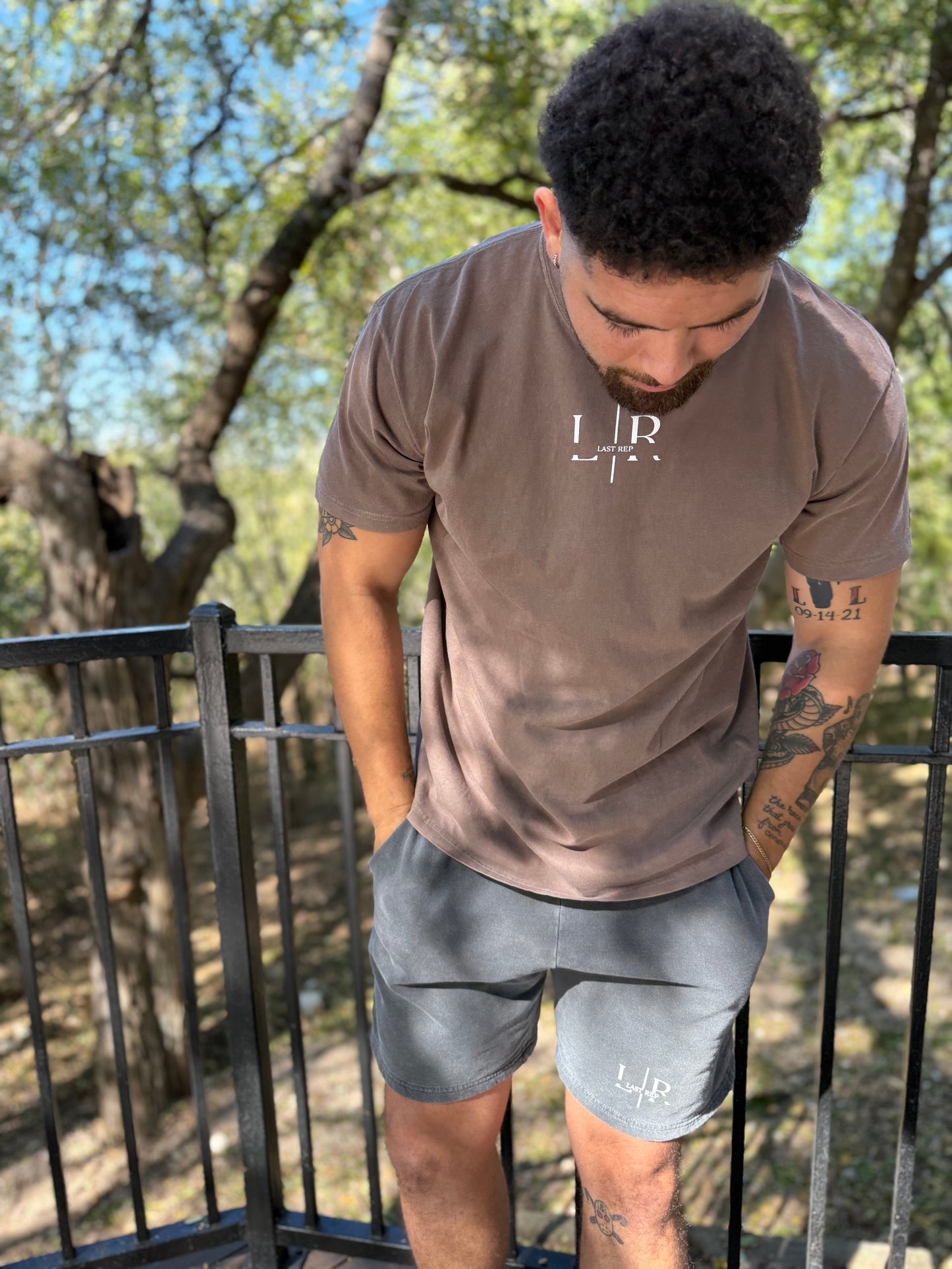 Man wearing a brown t-shirt and gray shorts with a logo outdoors.