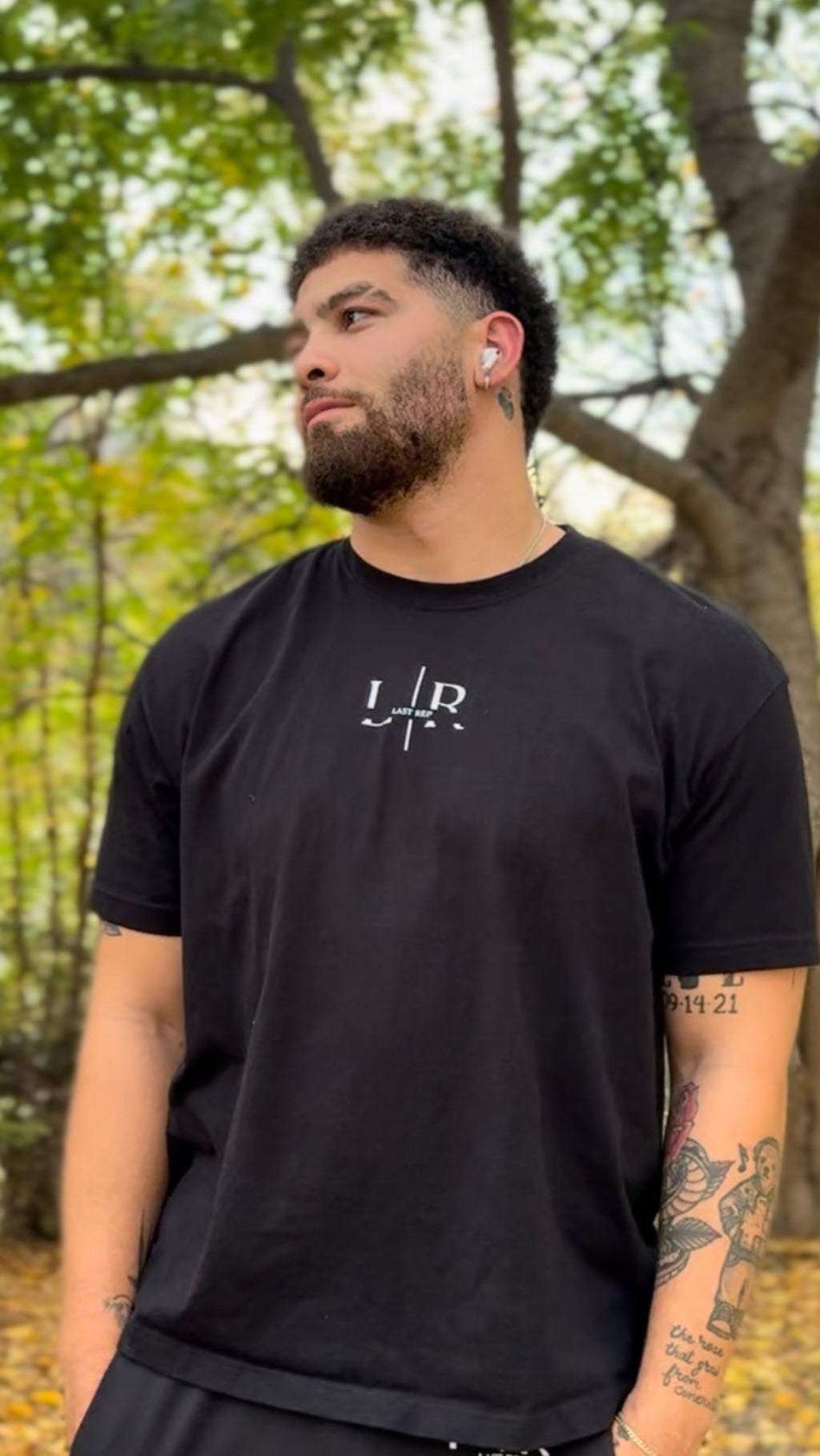 Man wearing a black t-shirt with a logo in an outdoor setting with trees and fallen leaves.