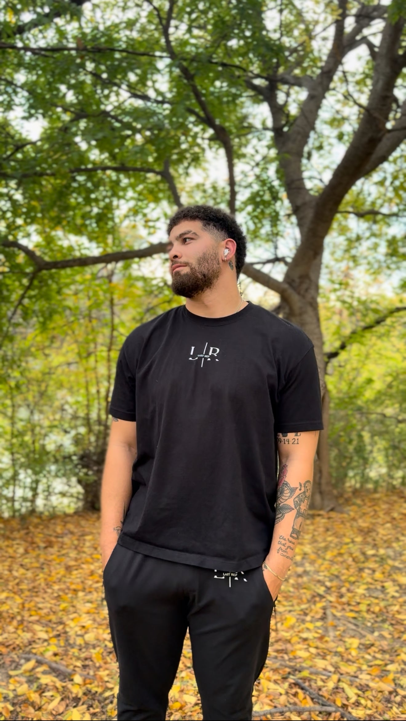 Man wearing a black t-shirt with a logo in an outdoor setting with trees and fallen leaves.