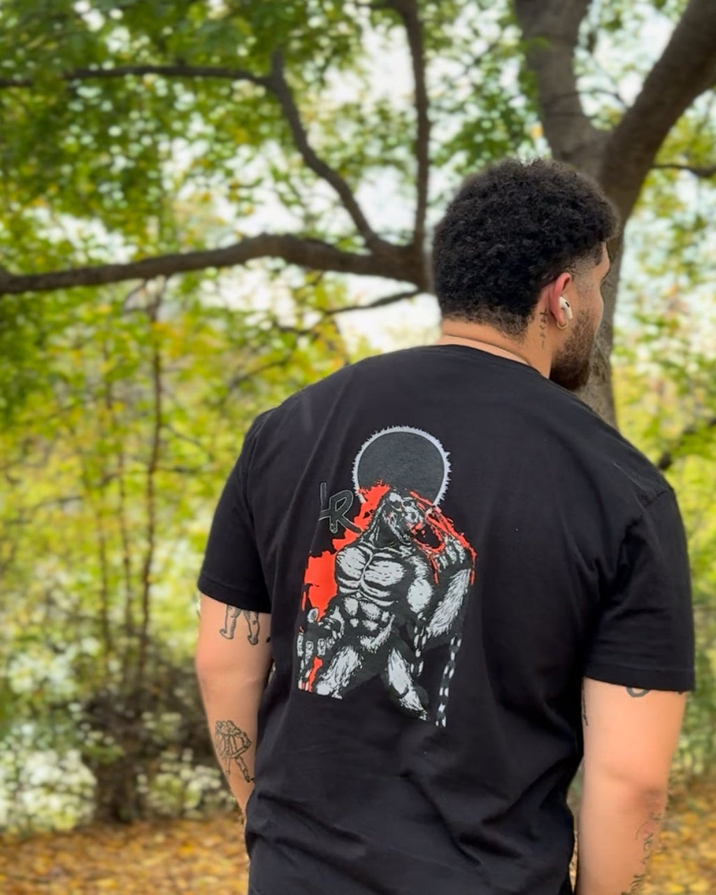 Person wearing a black t-shirt with a graphic design on the back, standing outdoors with trees in the background.