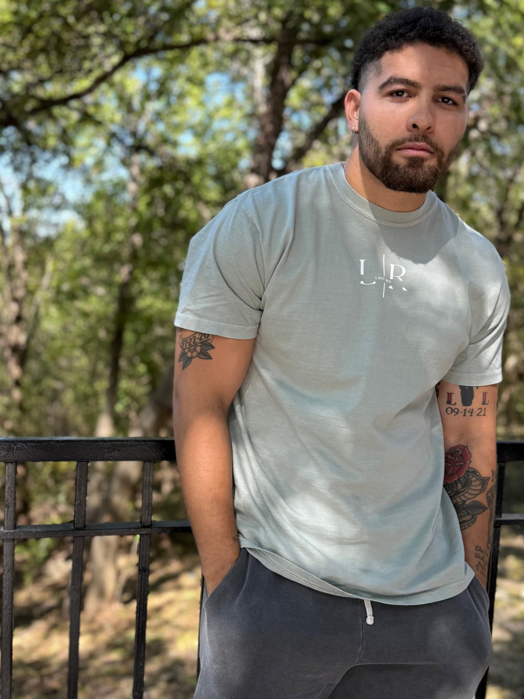 Man wearing a light green t-shirt and dark gray shorts with 'Last Rep' branding, standing outdoors with trees in the background.