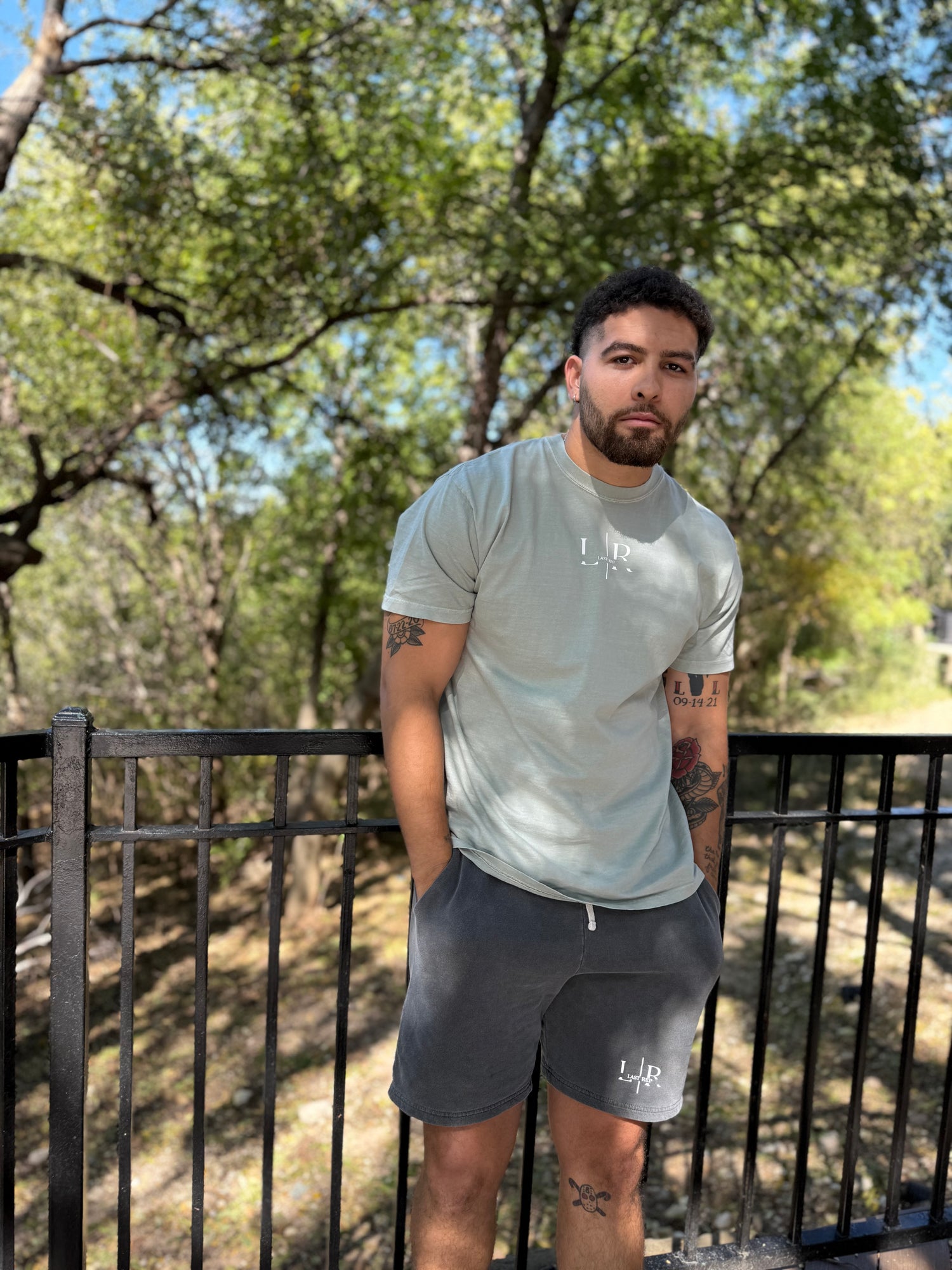 Man wearing a light green t-shirt and dark gray shorts with 'Last Rep' branding, standing outdoors with trees in the background.