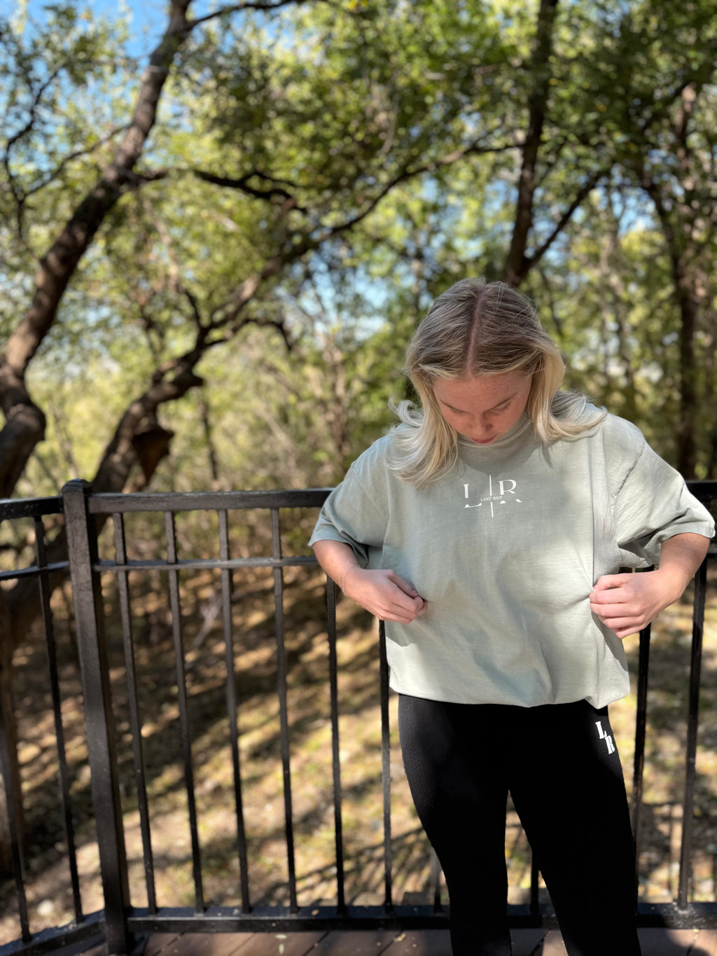 Person wearing a green shirt with text, standing outdoors on a wooden deck with trees in the background.