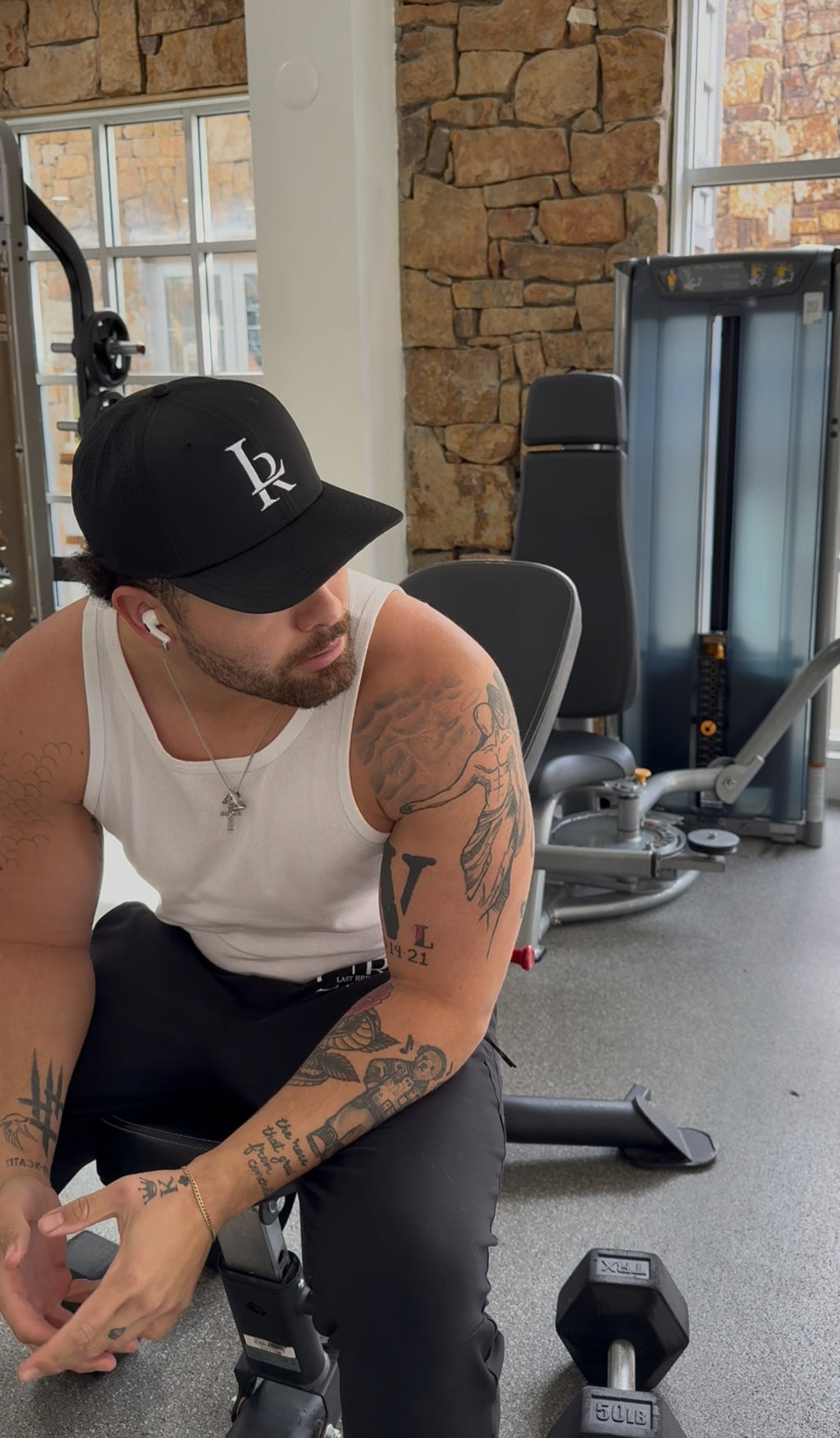 Man with tattoos in a gym setting wearing a cap and tank top.
