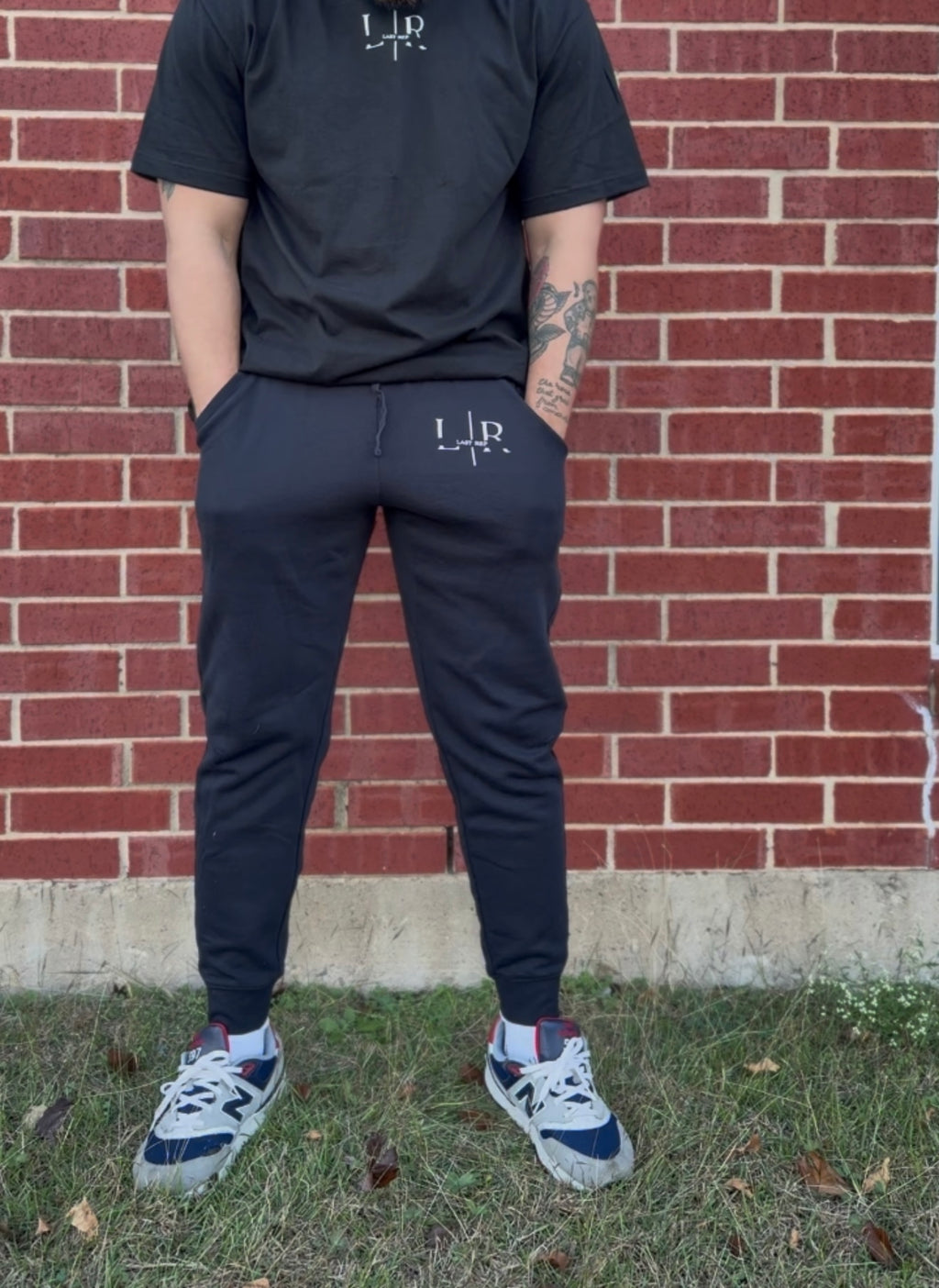 Person wearing black t-shirt and jogger pants with a logo, standing against a brick wall.