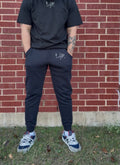 Person wearing black t-shirt and jogger pants with a logo, standing against a brick wall.