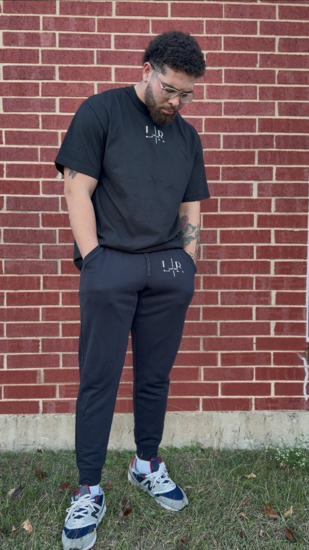 Man wearing black t-shirt and pants with a logo, standing against a brick wall.