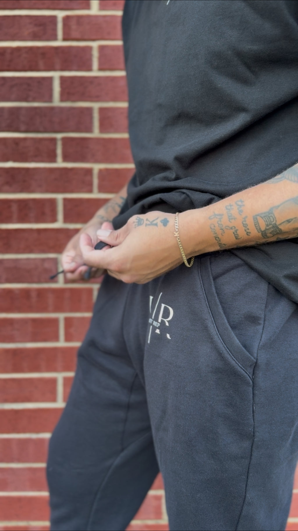 Person wearing dark pants with a logo, standing against a brick wall.