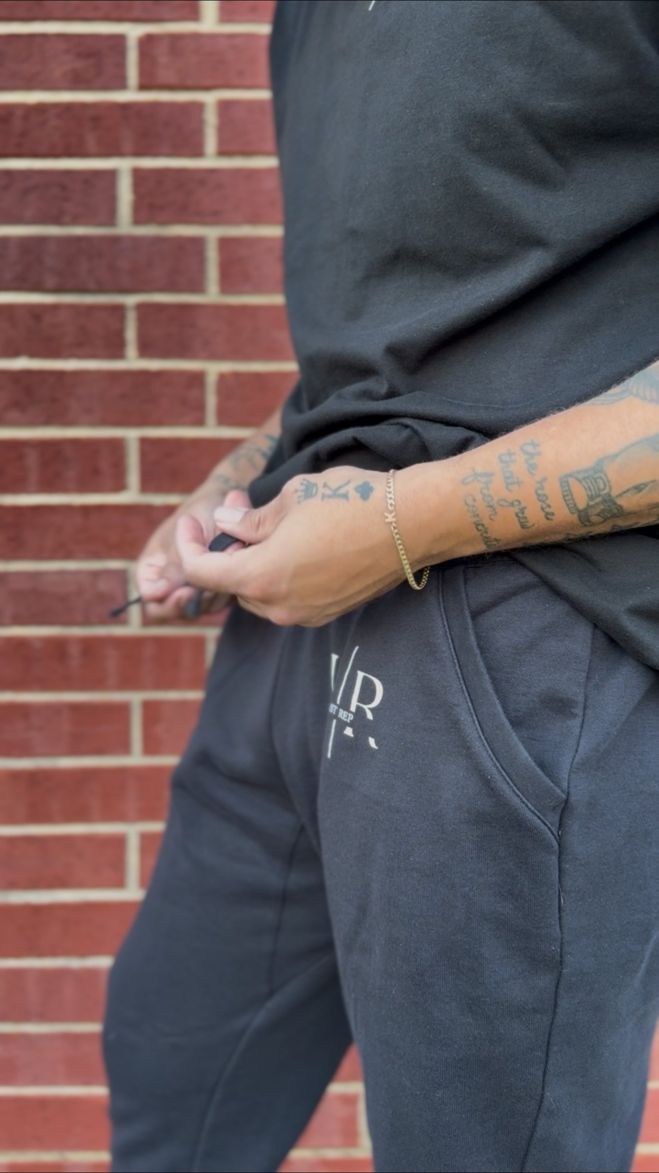 Person wearing dark pants with a logo, standing against a brick wall.