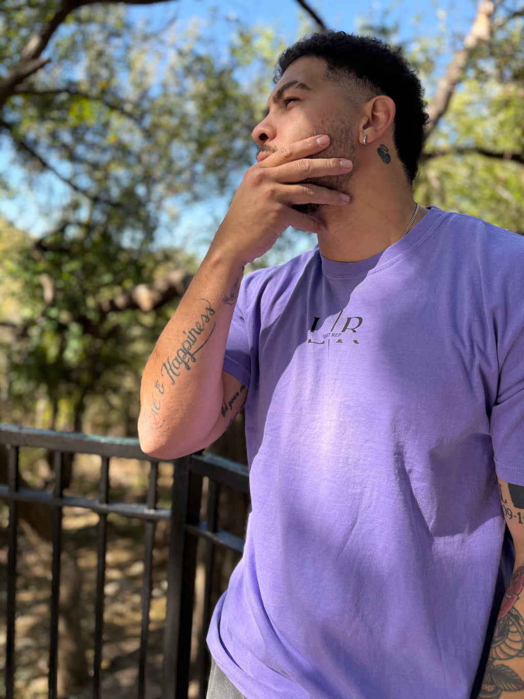 Man wearing a purple t-shirt with a logo, standing outdoors with trees in the background