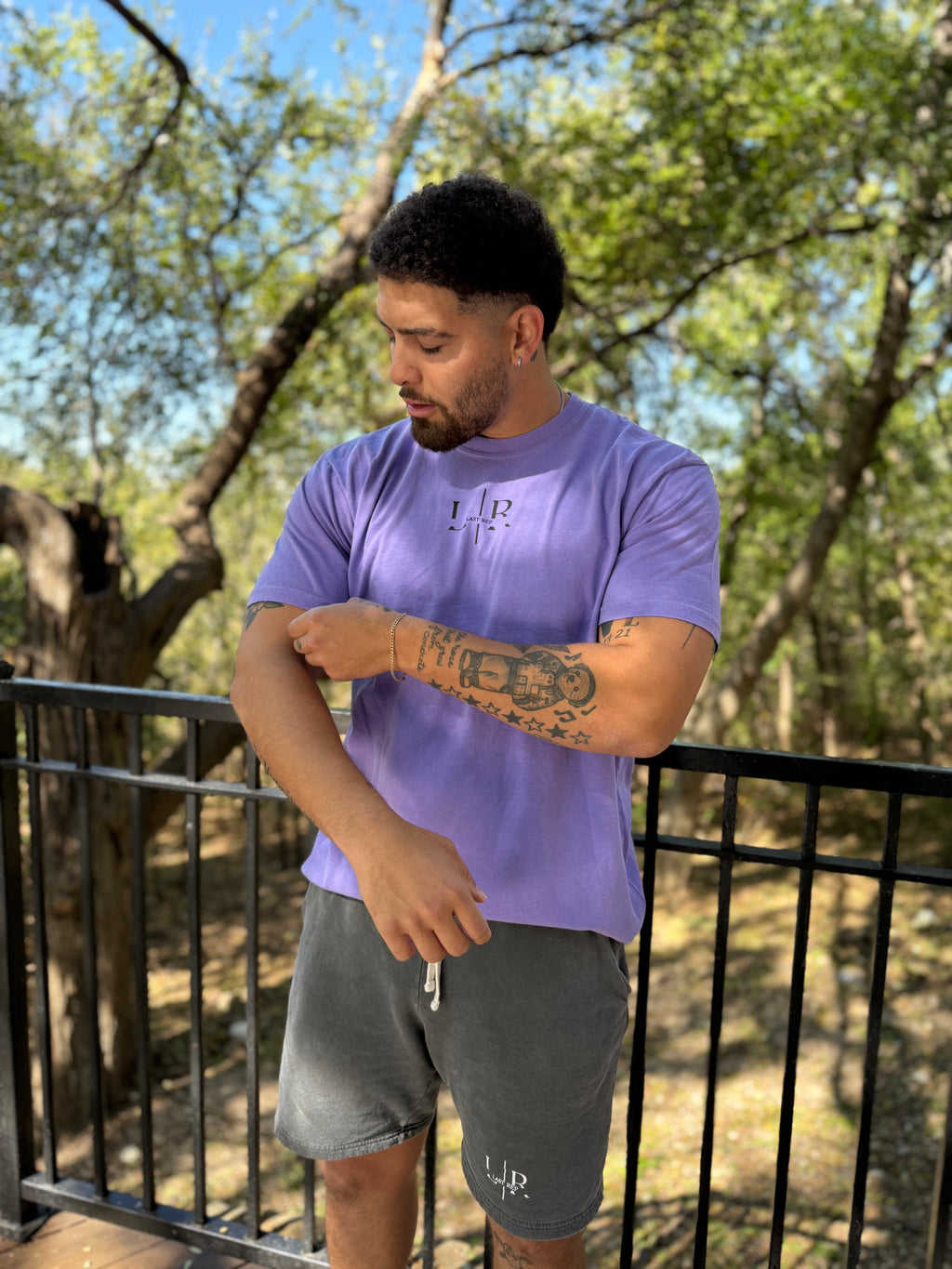 Man wearing a purple t-shirt with a logo, standing outdoors with trees in the background