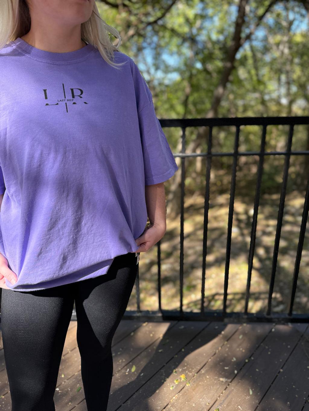 Person wearing a purple t-shirt with a logo on a wooden deck with trees in the background
