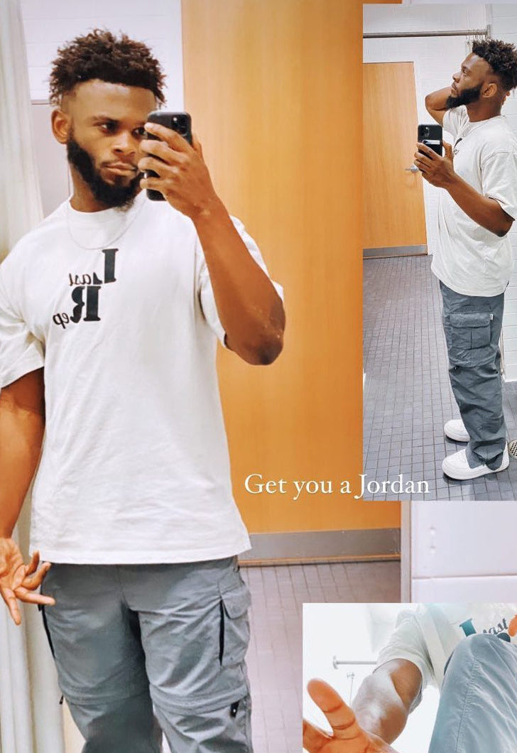 Man taking a mirror selfie wearing a tan Last Rep t-shirt and gray pants, with text 'Get you a Jordan' on an orange wall.
