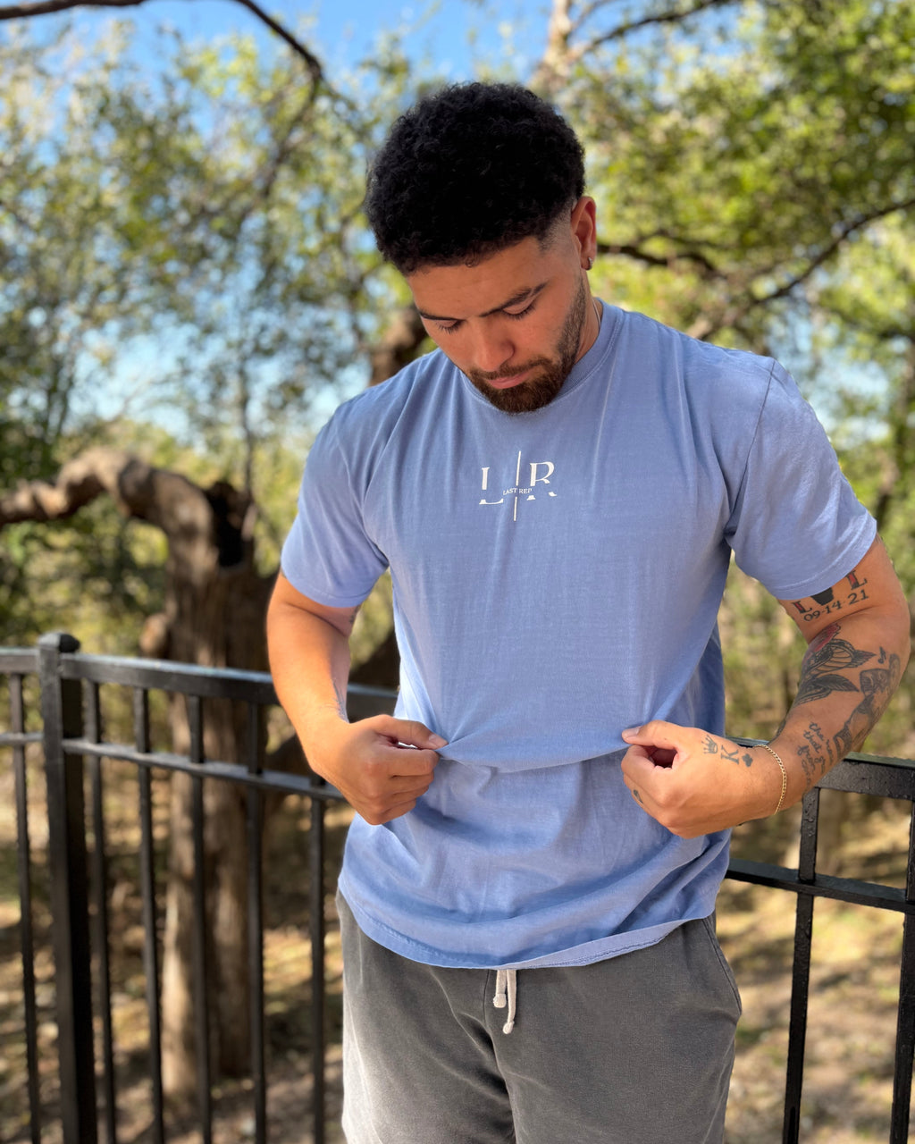 Man wearing a light blue t-shirt with a logo outdoors, surrounded by trees and animals.