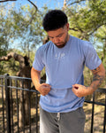 Man wearing a light blue t-shirt with a logo outdoors, surrounded by trees and animals.