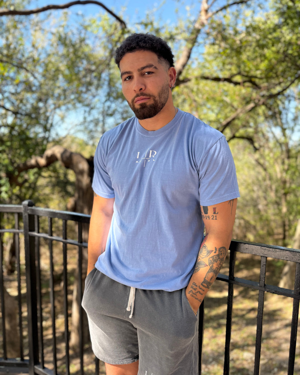 Man wearing a blue t-shirt and gray shorts standing outdoors with trees in the background