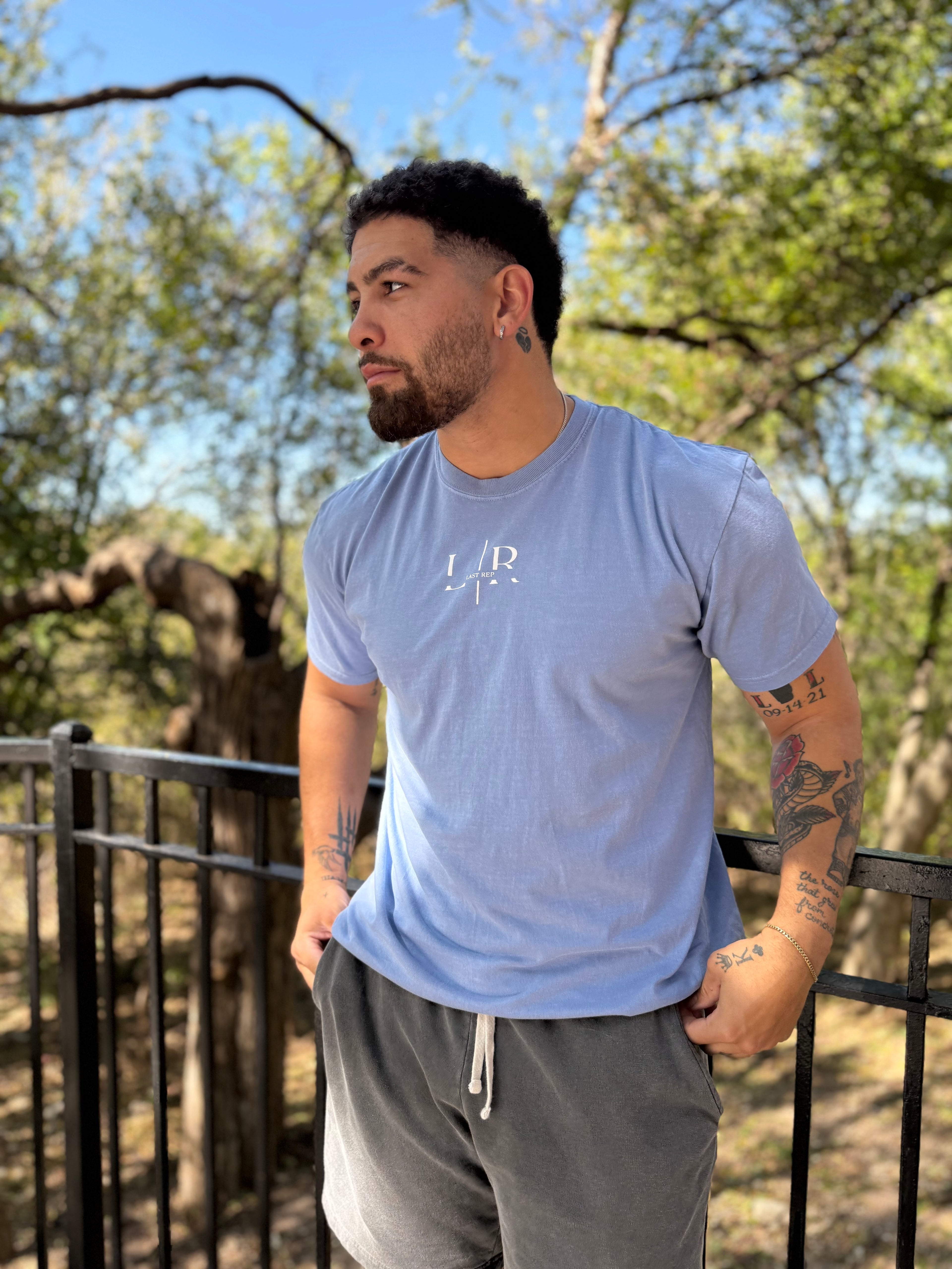 Man wearing a light blue t-shirt with a logo, standing outdoors with trees in the background