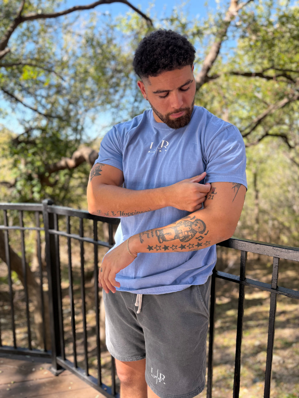 Man wearing a blue t-shirt and gray shorts with tattoos, standing outdoors.