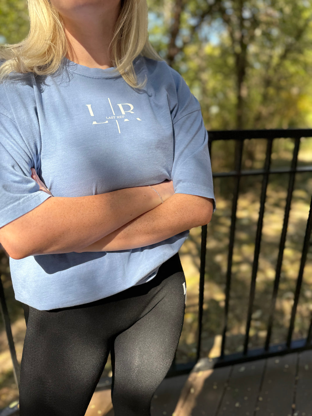 Person wearing a light blue t-shirt with a logo, standing outdoors with arms crossed.