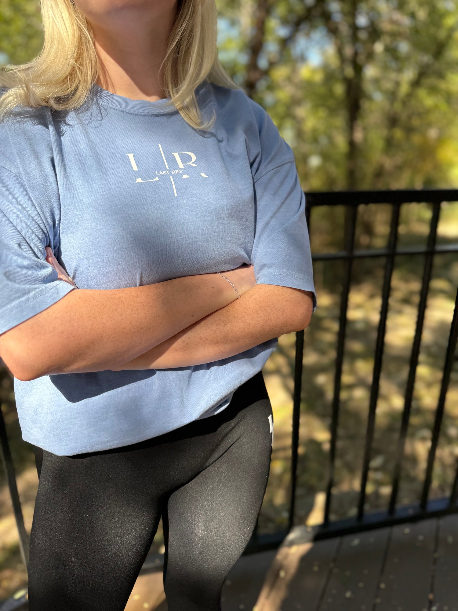 Person wearing a light blue t-shirt with a logo, standing outdoors with arms crossed.