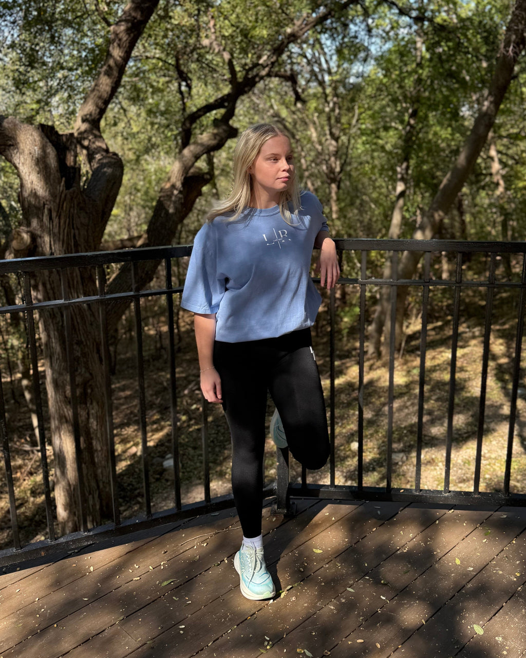 Person wearing a blue sweatshirt and black leggings standing on a wooden deck with trees in the background