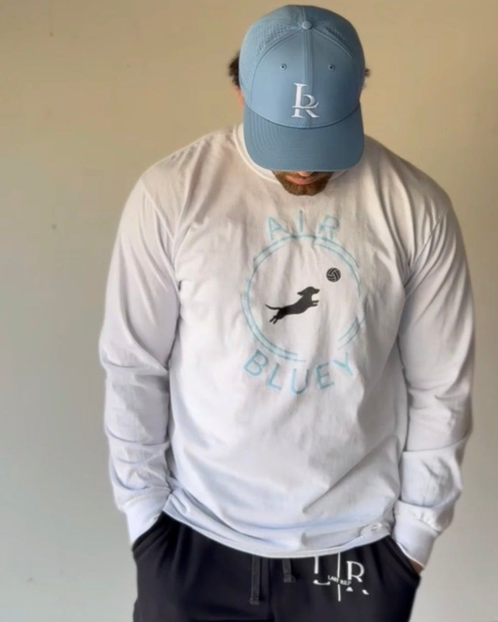 Person wearing a white long sleeve shirt with a logo and text, paired with a blue cap indoors.