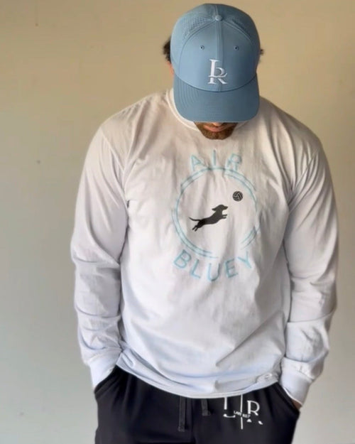 Person wearing a white long sleeve shirt with a logo and text, paired with a blue cap indoors.