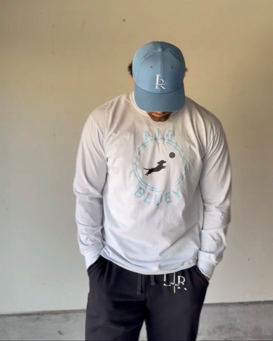 Person wearing a white long sleeve shirt with a logo and text, paired with a blue cap indoors.