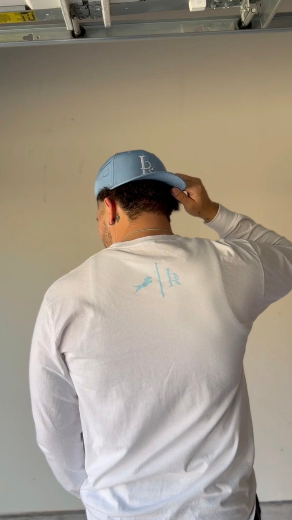 Person wearing a white long-sleeve shirt with a logo, adjusting a blue cap indoors.