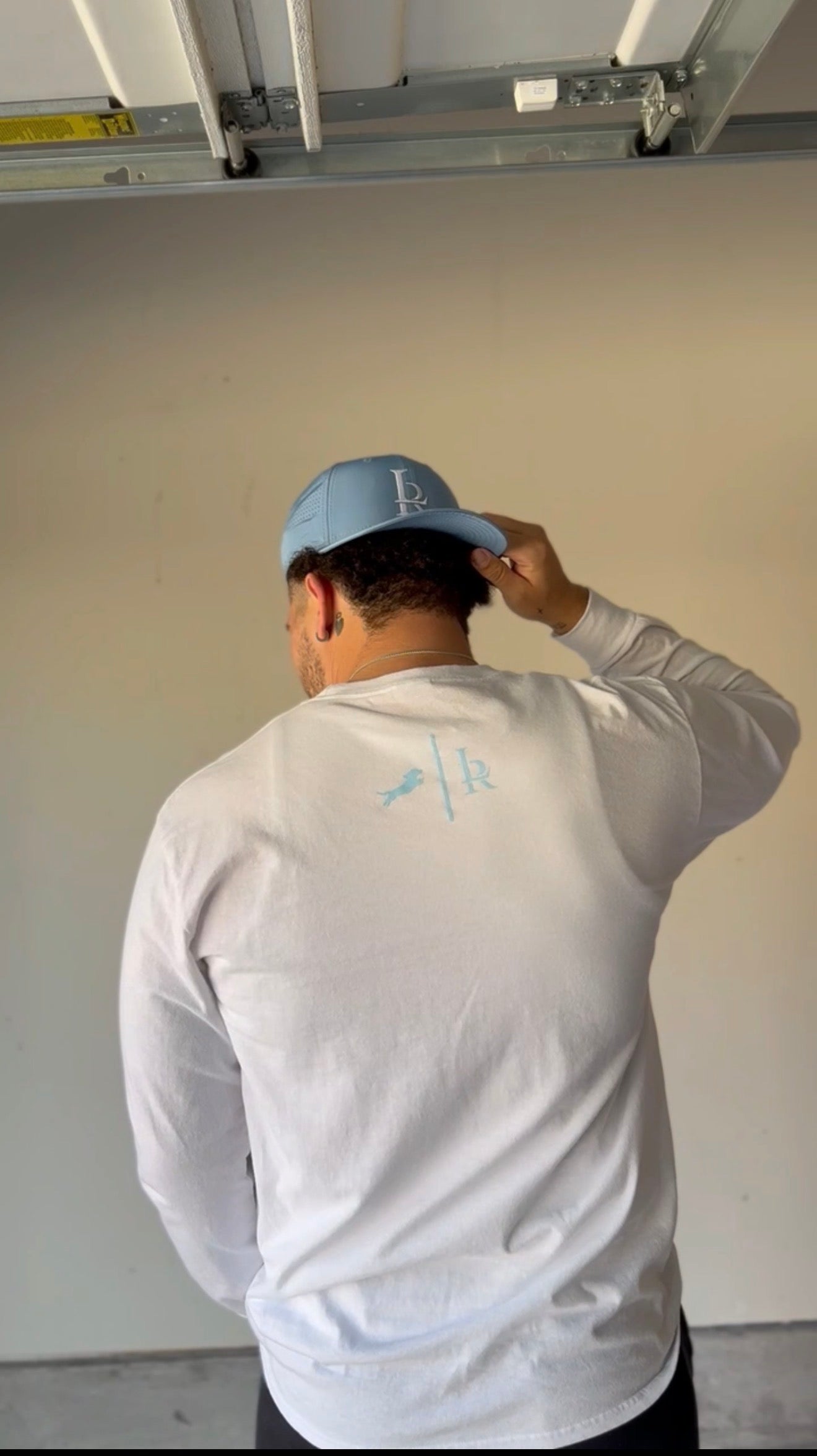 Person wearing a white long-sleeve shirt with a logo, adjusting a blue cap indoors.
