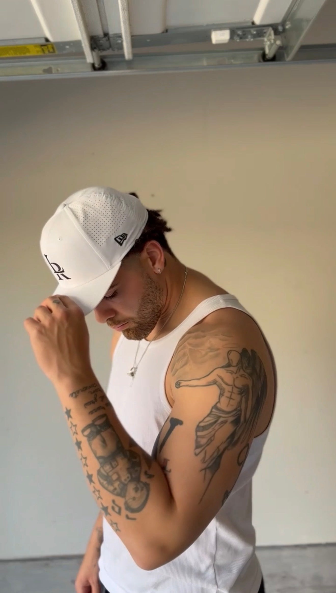 Man wearing a white cap and tank top with tattoos on his arms, standing in a garage.