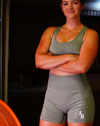 Woman in green sports bra and gray shorts standing with arms crossed against a dark background