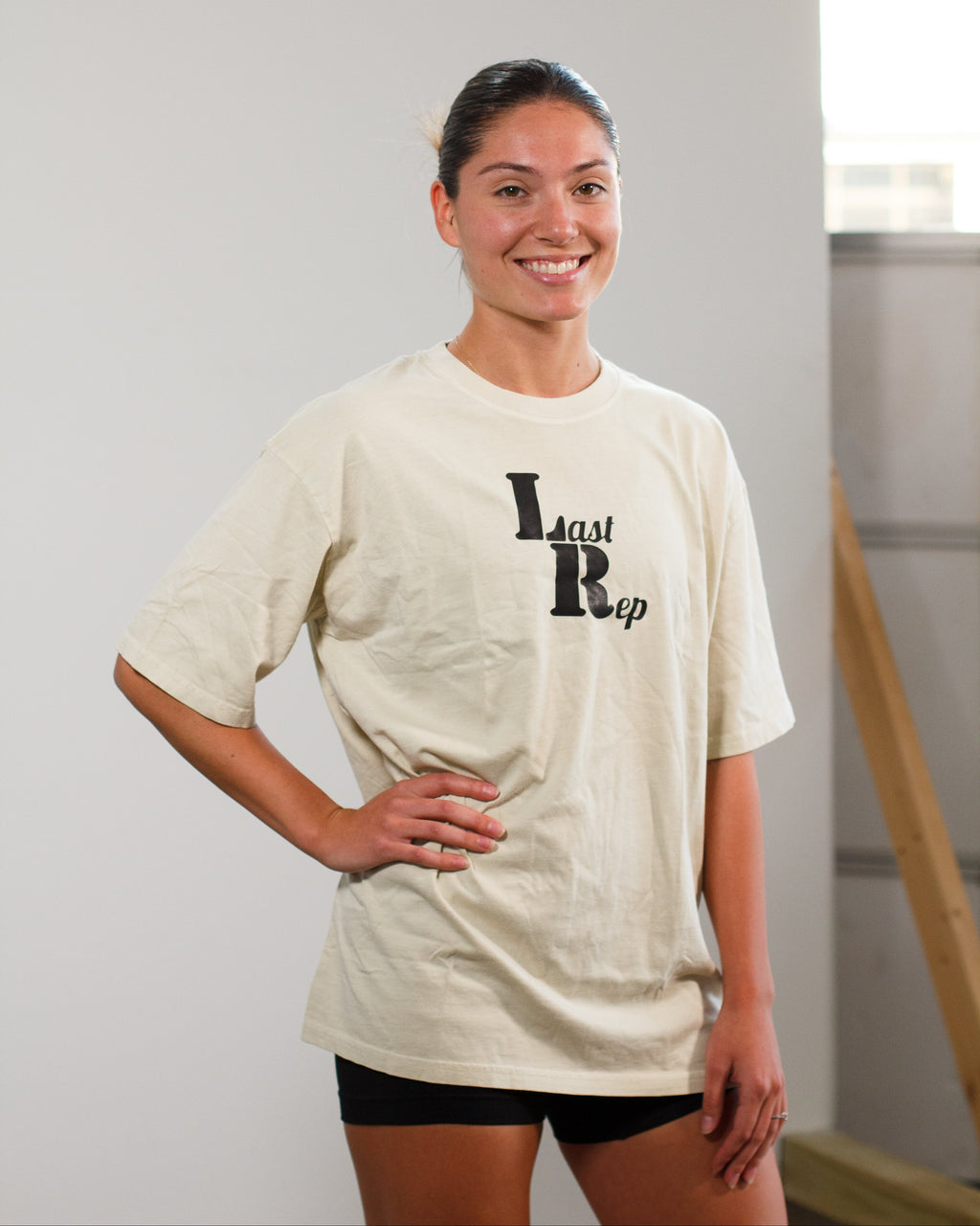 Woman wearing a beige t-shirt with Last Rep logo' text, standing indoors.